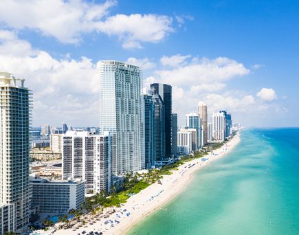 Blick auf Miami Beach. Links die hohen Hotels und Geschäftsgebäude, rechts daneben ein breiter weißer Sandstrand, daneben dann das türkisblaue Meer. Der Himmel strahlt hellblau.