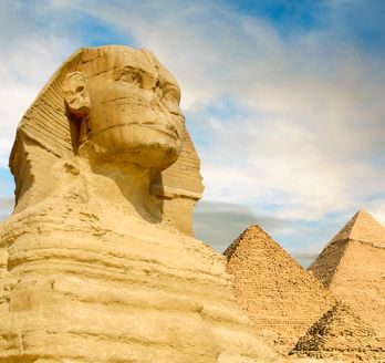Eine Nahaufnahme der Großen Sphinx von Gizeh, mit zwei der Pyramiden von Gizeh im Hintergrund unter einem bewölkten Himmel.
