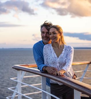Eine Frau steht an der Reling eines Schiffes und blickt auf den Sonnenuntergang über dem Meer.