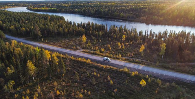 Camper Van fährt auf langer Straße in Schweden zwischen Wald und entlang eines Gewässers.