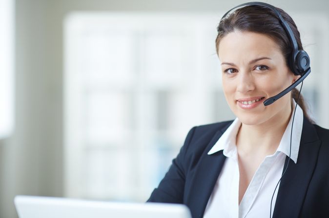  Eine Frau sitzt vor einem Computerbildschirm mit einem Headset und Mikrofon, was auf einen Kundenservice-Arbeitsplatz hindeutet. Sie blickt konzentriert auf den Bildschirm, während im Hintergrund weitere Arbeitsplätze unscharf zu sehen sind.