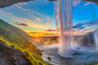 Blick aus einer Höhle, über welcher ein Wasserfall entspringt mit Gewässer unterhalb und grüner Landschaft bei Sonnenuntergang in Island
