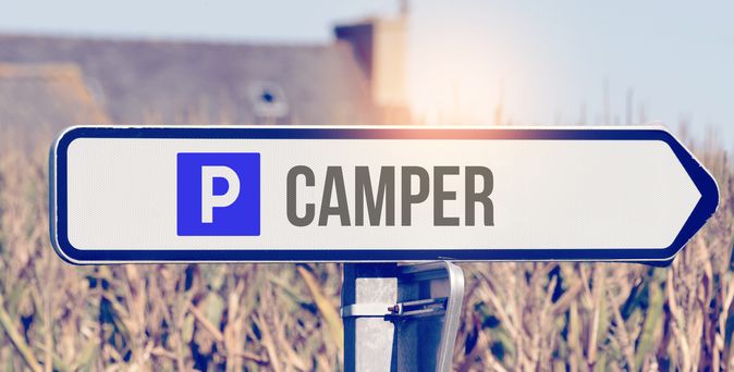 Blaues Schild mit weißem Pfeil und dem Symbol "P Camper" auf einem Schotterweg.