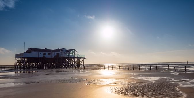 Ein ikonischer Holzpfahlbau in Sankt Peter-Ording steht am weiten Sandstrand des Wattenmeers, mit der Nordsee im Hintergrund und einem weiten Himmel darüber.