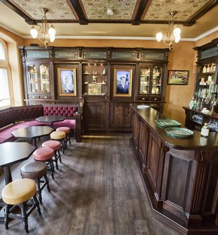 Ein traditioneller Pub-Innenraum mit einer langen, dunklen Holztheke, hohen Hockern mit gelben Polstern und einer reich verzierten Holzvertäfelung an den Wänden. Im Hintergrund sind auf Holzregalen Flaschen zu sehen.