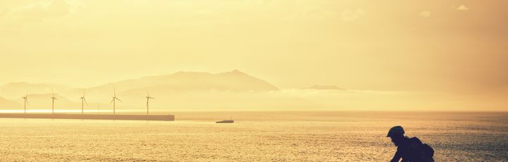 Eine Person fährt mit dem Fahrrad auf einer Straße, die am Meer entlangführt, während die untergehende Sonne den Himmel in warme Farben taucht. Die Szene wirkt ruhig und idyllisch.