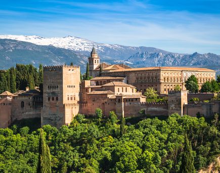 Blick auf den Palast Alhambra in Granada Spanien. Der Palast ist umgeben von Wald und im Hintergrund sind Berge mit vereinzelt Schnee auf den Spitzen zu sehen.