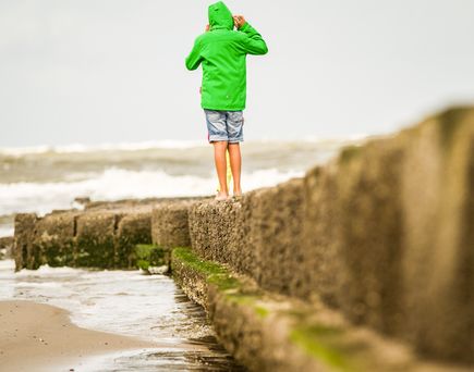 Ein Kind mit grüner Jacke und kurzer Hose spaziert auf einem Steinsteg am Strand.