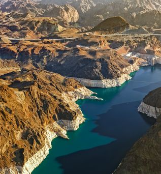 Eine Luftaufnahme des Hoover-Staudamms, der einen tiefblauen See in einer kargen, felsigen Schlucht staut. Die umliegenden Berge sind in Braun- und Rottönen gehalten, und eine große Bogenbrücke überspannt die Schlucht in der Ferne.