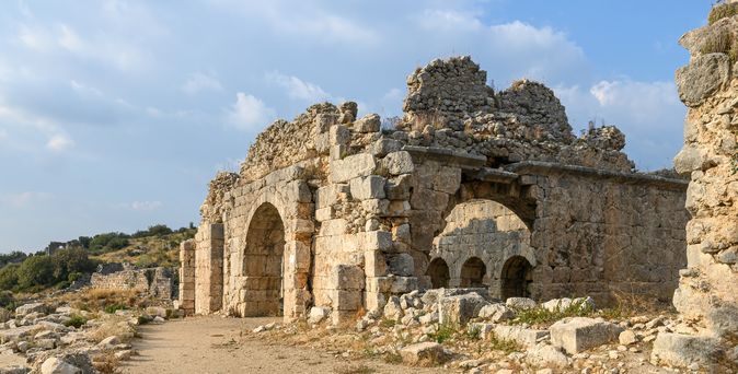Die Überreste der antiken Stadt Tlos in der Türkei, mit imposanten Steinruinen und einem Pfad unter einem bewölkten Himmel.