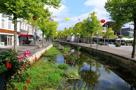 Zentrum von Heerenveen. Am Wasser, kleine Häuser und Cafes zu sehen, umgeben von Bäumen und Wiese. Bunte Ballons steigen.