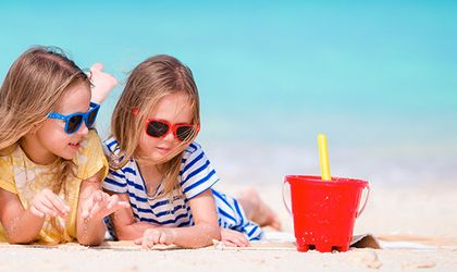 Zwei kleine Mädchen mit Sonnenbrillen spielen am Sandstrand mit bunten Eimern vor türkisblauem Meer.