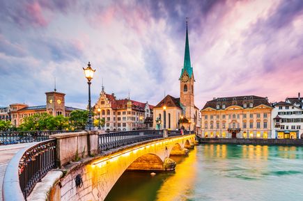 Eine kleine Brücke führt in die Stadt Zürich, umgeben von Wasser und kleinen Häuser. Das alles in der Dämmerung mit rotblauem Himmel.