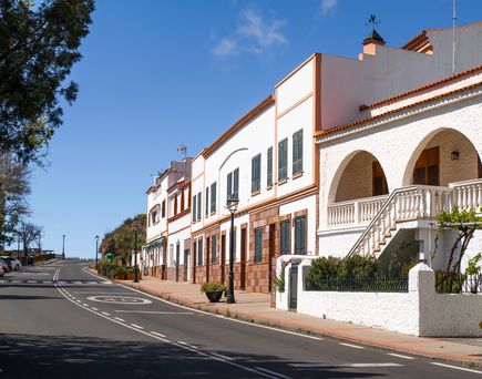 Bergdorf auf Gran Canaria. Schmale asphaltierte Straße, links einige bäume, rechts einige flache weiße Häuser. Blauer Himmel.