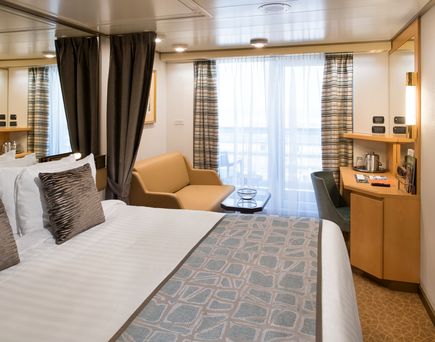 Blick in eine Verandakabine. Zuerst das große Doppelbett zu sehen, daher ein Sofa, rechts ein Schreibtisch und gegenüber eine große Balkon-Glastür für die Veranda.