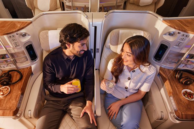 Paar stößt mit Getränken in nebeneinanderliegenden Business-Class-Sitzen an und lächelt sich an.