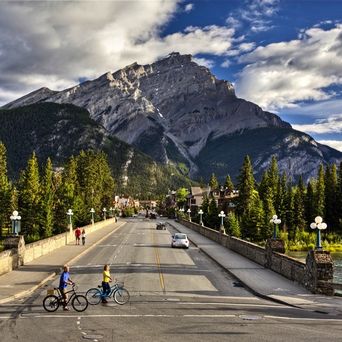 Majestätischer Blick auf den Cascade Mountain, der sich über der Stadt Banff in Alberta, Kanada, erhebt, mit den bewaldeten Ausläufern der Rocky Mountains und dem Bow River im Vordergrund, unter einem leicht bewölkten Himmel.