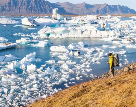 Trecking. Ein Mann mit gelber Windjacke steht am Ufer des gefrorenen Flusses. Dahinter Eisberge.