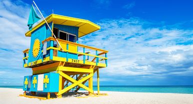 Ein blau-gelber Rettungsschwimmerturm am Sandstrand. Dahinter blaues Meer und blauer Himmel