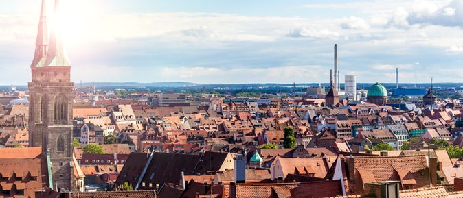 Panoramaaufnahme der Altstadt von Nürnberg mit markanten Kirchtürmen, roten Dächern und Industrieanlagen am Horizont.