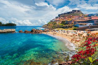 Sandstrand mit türkisblauem Wasser und blühenden Pflanzen. Am Stand liegen Personen zum Baden und im Hintergrund ist ein Dorf mit bunten Häusern in Italien
