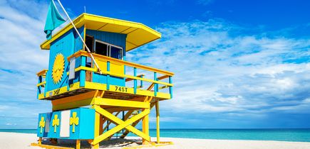 Ein blau-gelber Rettungsschwimmerturm am Sandstrand. Dahinter blaues Meer und blauer Himmel