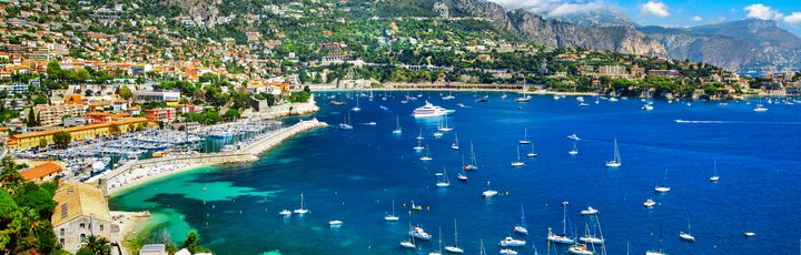 Eine Luftaufnahme einer Küstenstadt mit einer Bucht, vielen Booten und Gebäuden, umgeben von grünen Hügeln.