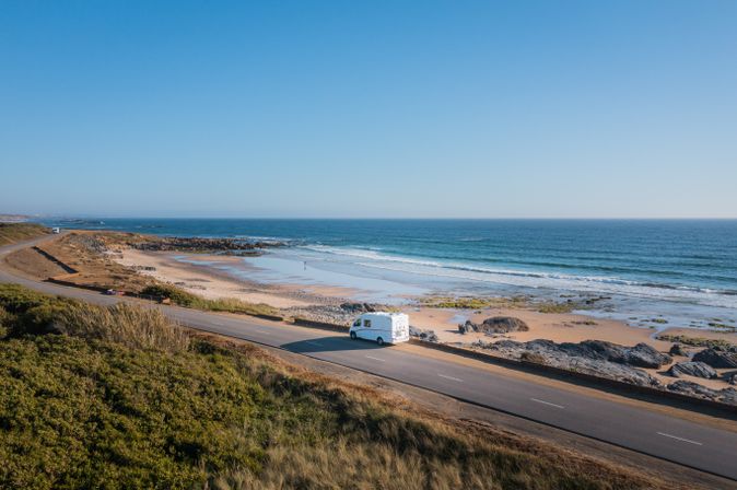 Wohnmobil fährt auf Küstenstraße entlang einem Strand mit kleinen Felsen und blauem Meer mit Felsen in Irland