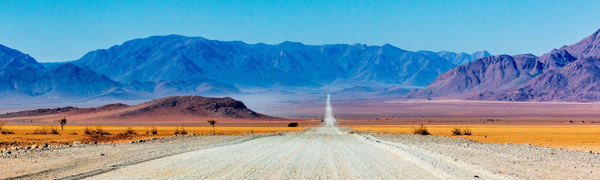Eine lange, gerade Straße erstreckt sich durch die beeindruckende Wüstenlandschaft des Namib-Naukluft Nationalparks in der Namib-Wüste, umgeben von roten Sanddünen und einem weiten, blauen Himmel.