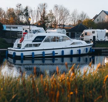 Ein kleines, weißes Motorboot mit dem Schriftzug "CARRICKCRAFT" und blauen Akzenten, das auf einem ruhigen Fluss oder See fährt. Im Hintergrund ist eine bewaldete Uferlandschaft mit weiteren Booten zu sehen.