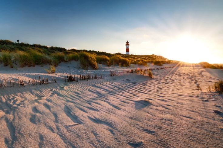 Leuchtturm auf einer Düne im Sonnenuntergang mit langen Schatten auf dem fein strukturierten Sand.