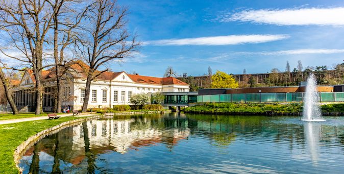 Ein eleganter Kursaal im Kurpark Bad Kösen, umgeben von gepflegten Grünflächen und Bäumen, unter einem klaren blauen Himmel.