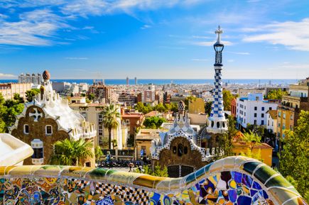 Park Güell in Barcelona in Spanien, mit den charakteristischen bunten Mosaikbänken im Vordergrund. Im Hintergrund erstreckt sich die Stadt Barcelona bis zum Mittelmeer unter einem klaren blauen Himmel.