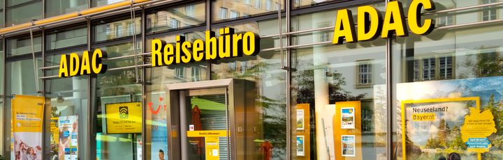 ADAC Reisebüro mit gläserner Fassade und gelber Beschriftung in einer Innenstadtlage, Schaufenster mit Urlaubsangeboten.