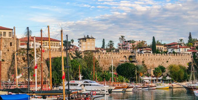 Der historische Hafen von Antalya, Türkei, bei Sonnenuntergang, mit zahlreichen Segelbooten und Yachten, die am Kai liegen, und der Silhouette der Altstadt im Hintergrund.