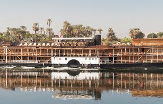 Ein historisches Dampfschiff, die SS Sudan, fährt auf dem Nil, umgeben von üppiger grüner Vegetation und dem ruhigen Wasser.