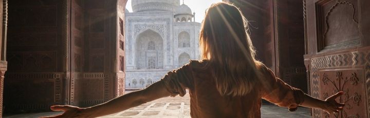 Frau mit ausgebreiteten Armen steht ergriffen im Eingangstor und blickt auf das leuchtende Taj Mahal im Morgenlicht.
