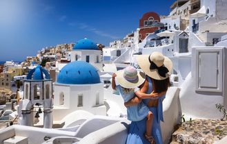 Eine Mutter und ihre Tochter stehen auf einem Balkon und blicken auf eine weiße Stadt und das Meer.