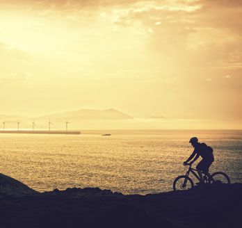 Eine Person fährt mit dem Fahrrad auf einer Straße, die am Meer entlangführt, während die untergehende Sonne den Himmel in warme Farben taucht. Die Szene wirkt ruhig und idyllisch.