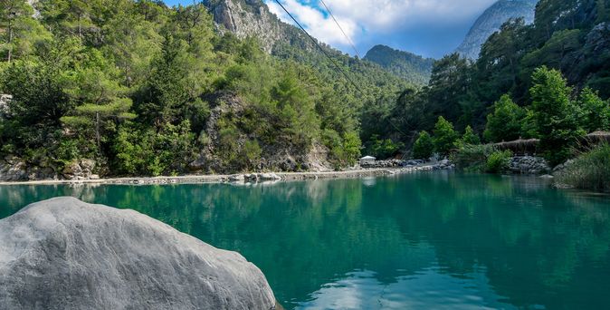 Blick über einen türkisblauen See, von Felsen und Wald umgeben. Strahlender Himmel