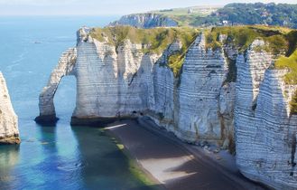 Eine felsige Küstenlinie mit hohen, weißen Klippen, die ins Meer ragen. Ein Segelboot schwimmt im türkisblauen Wasser.