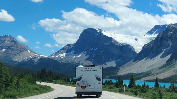 Eine atemberaubende Panoramaansicht des Icefields Parkway im Banff Nationalpark, Kanada, mit einer Straße, die sich durch eine majestätische Berglandschaft schlängelt, umgeben von schneebedeckten Gipfeln und einem weiten blauen Himmel.