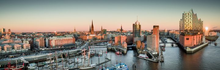 Ein Panorama von Hamburg bei Sonnenuntergang zeigt die Elbphilharmonie, den Hafen mit Schiffen und die Skyline der Stadt.