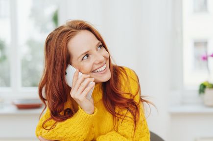 Eine rothaarige Frau in einem gelben Pullover lächelt, während sie ein Smartphone an ihr Ohr hält, offenbar im Gespräch.