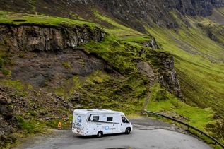Eine dramatische Landschaftsansicht des Quiraing auf der Isle of Skye, Schottland, mit grünen Hügeln, schroffen Felsen und einem Parkplatz im Vordergrund unter einem dramatischen Himmel.