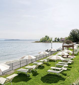 Grüne Liegewiese mit Sonnenschirmen und Liegen direkt am Ufer eines Sees mit Kiesstrand und klarer Sicht auf das Wasser.