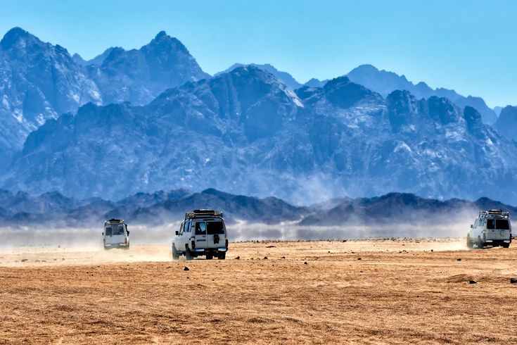 Mehrere weiße Geländewagen fahren im Konvoi durch die staubige Wüste vor einer eindrucksvollen, bläulichen Bergkette.