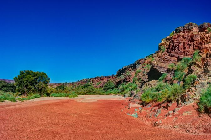  Ein unbefestigter Weg schlängelt sich durch eine trockene, felsige Landschaft, die von spärlicher Vegetation bewachsen ist, unter einem weiten, klaren Himmel.