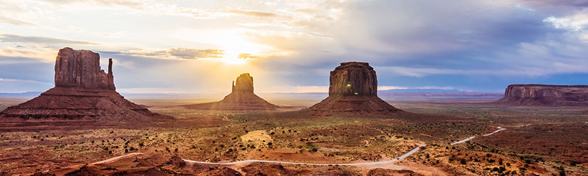 Ein Panorama des Monument Valley, Utah, USA, bei Sonnenuntergang oder Sonnenaufgang, mit den charakteristischen Buttes und Mesas, die in warmen Farben leuchten, und dramatischen Wolken am Himmel, durch die Sonnenstrahlen brechen.