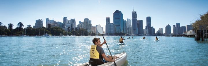 Panoramablick auf den Brisbane River, wo mehrere Personen in Kajaks paddeln, mit der beeindruckenden Kulisse der Hochhäuser von Brisbane im Hintergrund.